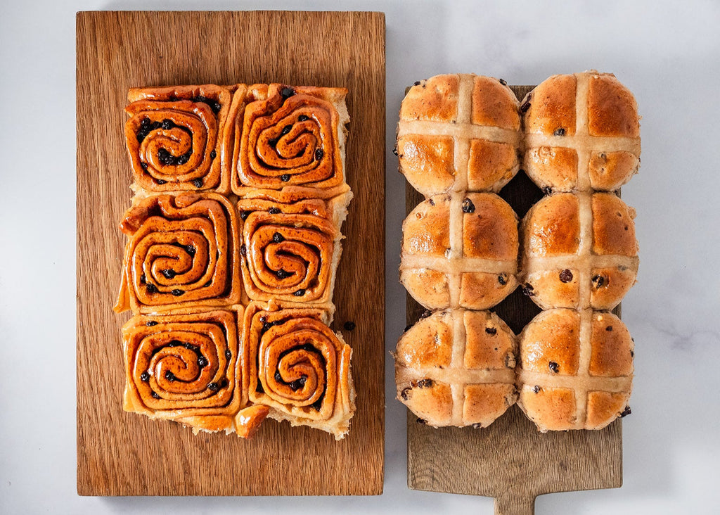 Six freshly baked Chelsea buns sit next to six freshly baked hot cross buns. They are on two rustic wooden boards.